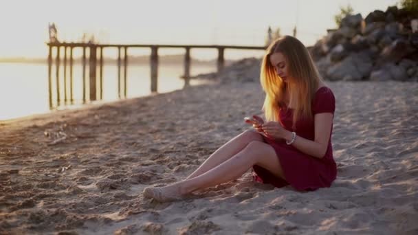 Смартфон жінка в червоній сукні смс-повідомлення, використовуючи додаток на смартфоні на заході сонця на пляжі . — стокове відео