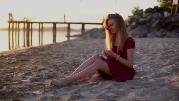 ビーチの夕日でスマート フォンのアプリを使って赤いドレスの sms テキスト メッセージのスマート フォン女性. — ストック動画