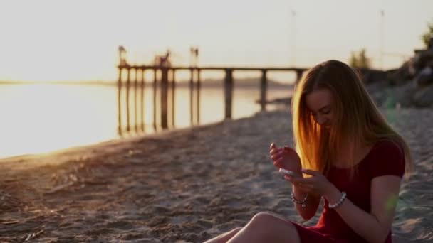 Smartphone-Frau im roten Kleid schreibt SMS per App auf Smartphone bei Sonnenuntergang am Strand. — Stockvideo