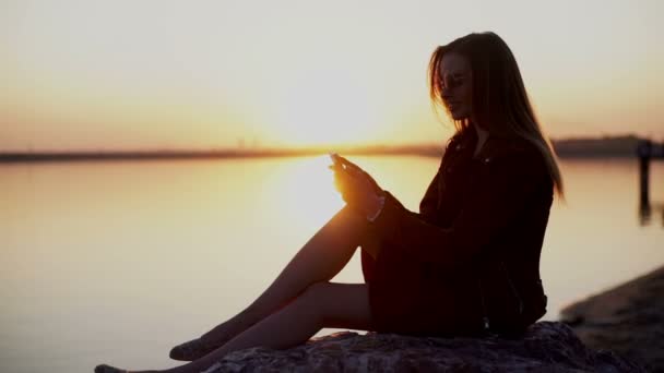 Смартфон жінка в червоній сукні смс-повідомлення, використовуючи додаток на смартфоні на заході сонця на пляжі . — стокове відео