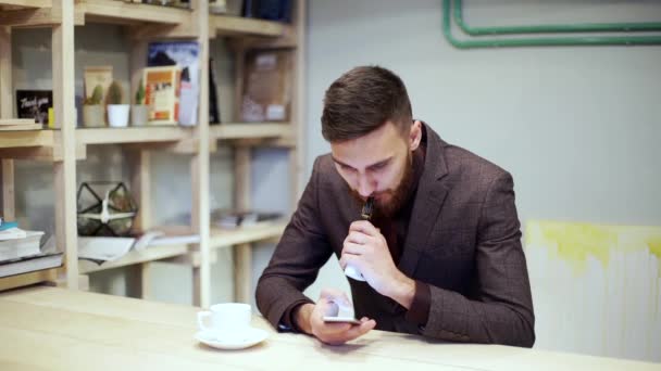 Barbudo hombre de traje está fumando vapor de cigarrillo electrónico, charlando por teléfono inteligente — Vídeos de Stock