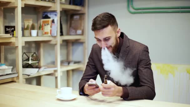 Barbudo hombre de traje está fumando vapor de cigarrillo electrónico, charlando por teléfono inteligente — Vídeo de stock