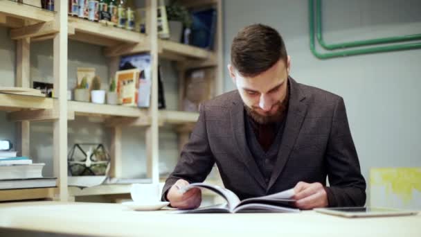 Jonge, bebaarde man is het lezen van tijdschrift in café, drinken koffie — Stockvideo