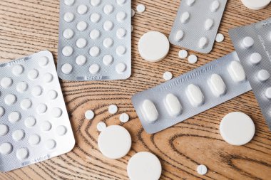 Beyaz yuvarlak haplar ve alüminyum paketlerde farklı boyutlarda oval haplar ve ahşap bir masaya saçılmış.