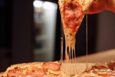 Eritilmiş peynir bir dilim pizzadan esniyor. Salamlı, mısırlı ve biberli bir dilim pizza yiyorum. Restoranda ahşap bir masada pizza.