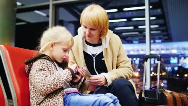 Девушка 5-6 лет, используйте свой телефон в терминале аэропорта, рядом с ее матерью сидит — стоковое видео