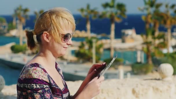Ein Tourist mit Sonnenbrille genießt das Tablet vor dem Hintergrund einer tropischen Landschaft — Stockvideo