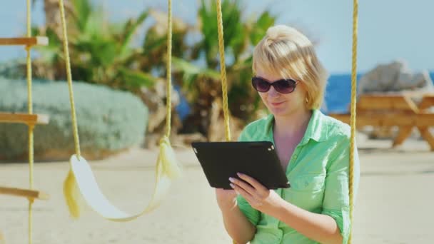 Ένα θηλυκό τουριστών απολαμβάνει το tablet, κάθεται σε μια κούνια — Αρχείο Βίντεο