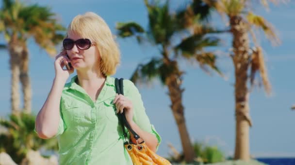Женщина-туристка в солнечных очках разговаривает по телефону на фоне неба и пальм — стоковое видео