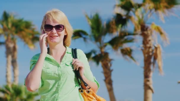 Turista feliz fala por telefone em um fundo de céu azul e palmeiras — Vídeo de Stock