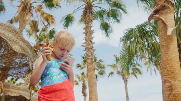 滑稽的女孩吃一根香蕉，在智能手机上播放 — 图库视频影像