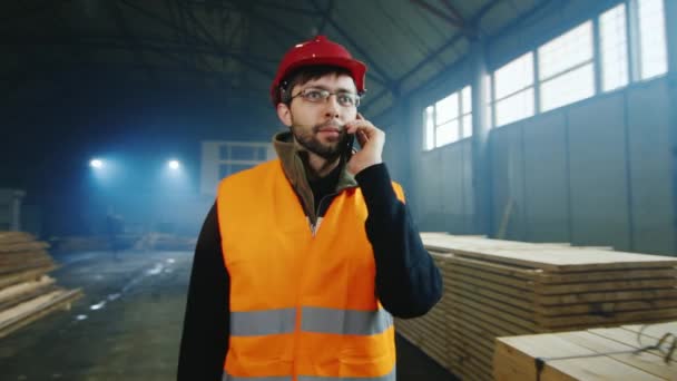 Steadicam geschoten: Builder in een helm en een heldere vest gaat over een magazijn van bouwmaterialen, praten over de telefoon — Stockvideo