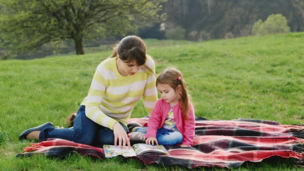 Una joven atractiva lee un libro, una niña pequeña. Sentado en el césped en el parque — Vídeo de stock