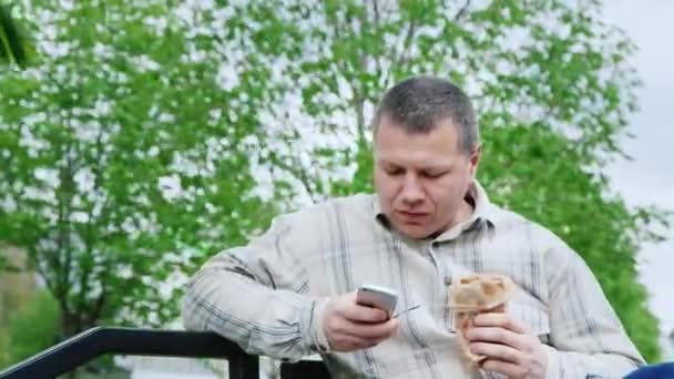 Человек в парке ест нездоровую пищу, пользуется твоим смартфоном — стоковое видео