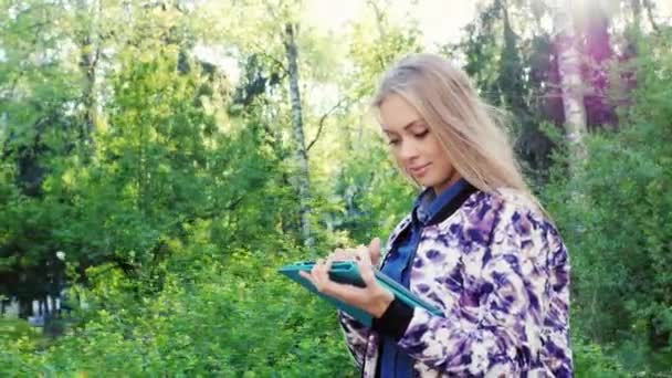 Mujer joven y atractiva caminando con una tableta en el parque de la ciudad — Vídeos de Stock