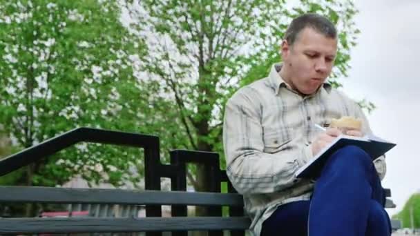Hombre comiendo comida chatarra en un banco del parque, escribe algo en un cuaderno — Vídeos de Stock