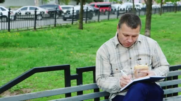 Un homme mange de la restauration rapide sur un banc de parc, écrit quelque chose dans un carnet — Video