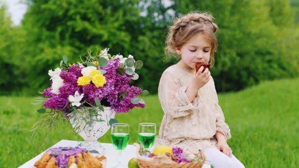 Vicces lány 4 év enni tortát egy piknik. Ő háttér zöld gyep édességek asztal körül — Stock videók