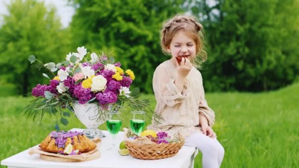 お菓子をテーブルの上に座って笑って面白い女の子。ピクニックにケーキを食べる。背景の緑の芝生で — ストック動画