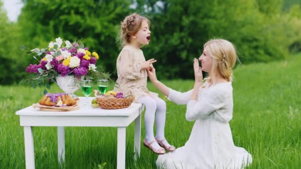 Μαμά παίζει με την κόρη της σε ένα πικνίκ. Κορίτσι που κάθεται σ ' ένα άσπρο τραπέζι με γλυκά στο παρασκήνιο και καταπράσινο γκαζόν — Αρχείο Βίντεο