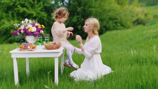 Девочка 4 лет играет со своей матерью - кормит ее кексом. Она сидит на столе со сладостями, на заднем плане зеленый газон — стоковое видео