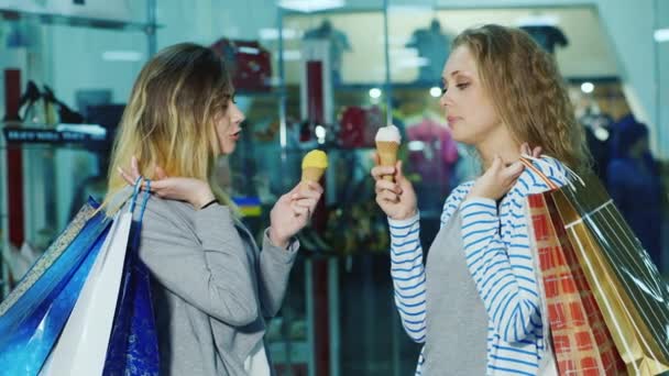 Dos amigos comiendo helado. De pie con bolsas de compras en una tienda en el fondo de vitrinas de vidrio — Vídeos de Stock