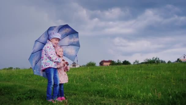 Två flickor gömmer sig från regnet under ett paraply. Mot bakgrund av en stormig himmel och en stark vind. — Stockvideo