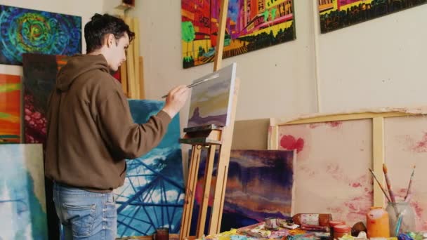 Jeune beau peintre travaillant dans l'atelier. Dessine un paysage rural américain typique — Video