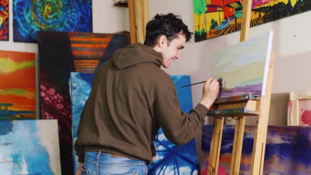 The young artist with a smile on his face paints a picture in the studio — Stock Video