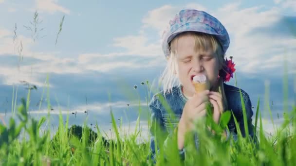 Grappig meisje buitenshuis eten van ijs. Hij zit in het groene gras tegen de hemel met prachtige wolken — Stockvideo