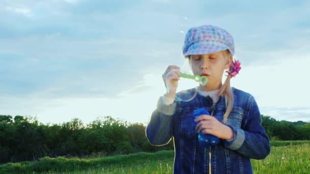 Dívku 5 let vyfukuje bubliny. Je to na zelené louce, slunce svítí a poskytuje pohled — Stock video