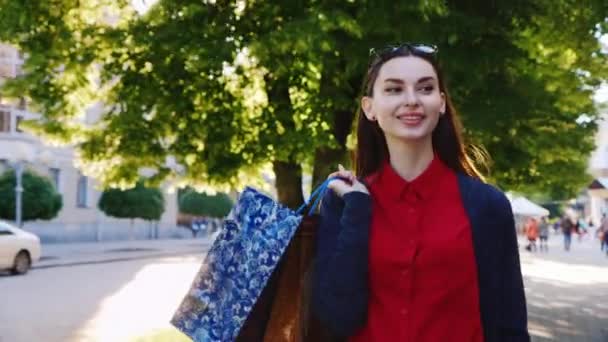 Alışveriş çantaları ile kırmızı elbiseli şık kadın Dyal şehirde dolaşıyor. Güneş parlıyor, çerçevede güzel yansımalar. — Stok video