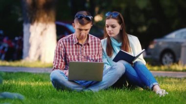 Parkta çimlerin üzerinde oturan bir erkek ve bir kadının arkadaşları, dizüstü bilgisayarın tadını çıkarıyorlar. Arka planda, insanlar yürümek