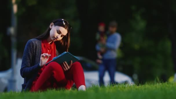 Stílusos nő ül a füvön a parkban, élvezve a tablettát. A háttérben az emberek sétálnak — Stock videók