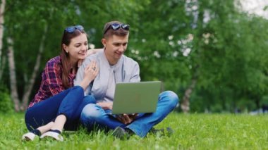 Parkta dinlenen, çimlerin üzerinde oturan ve dizüstü bilgisayarın keyfini çıkaran çekici çift