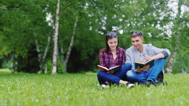 Unge man och kvinna läser en bok i parken. De sitter på en grön gräsmatta — Stockvideo