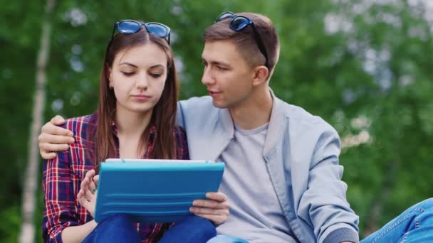 Joven chica y un chico utiliza una tableta en el parque, chico abraza chica — Vídeo de stock