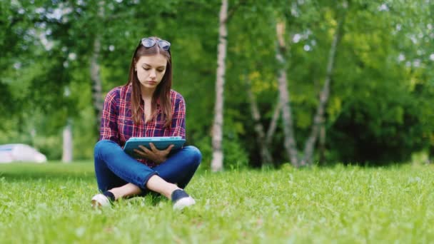 Linda mujer joven disfrutando de una tableta en el parque. Sentado en la hierba — Vídeo de stock