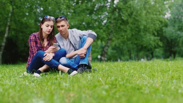 Друзья мужчины и женщины наслаждаются смартфоном, сидя на траве в парке — стоковое видео