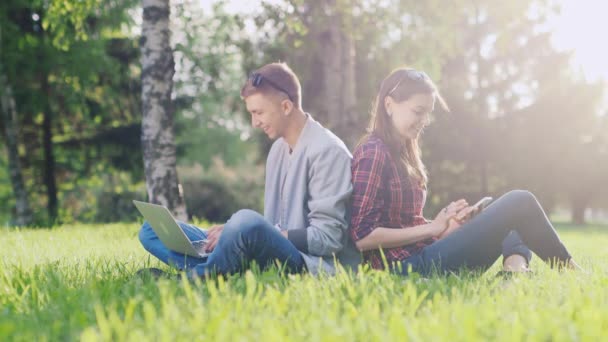 Los estudiantes que estudian en el parque - utilizar un ordenador portátil y un teléfono inteligente. Se sientan en la hierba verde — Vídeo de stock