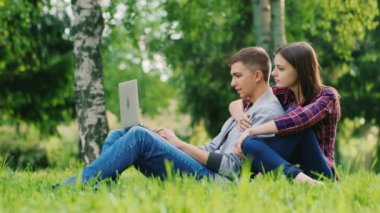 Laptop ile çalışma çimen parkta oturan bir çekici genç bir çift. Kız arkasındaki adam kucaklayan