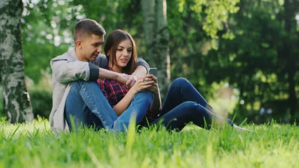 Mężczyzna i Kobieta, przytulanie, siedząc na trawie w parku. Patrząc na ekran telefonu komórkowego. Komunikować się, uśmiechając się — Wideo stockowe