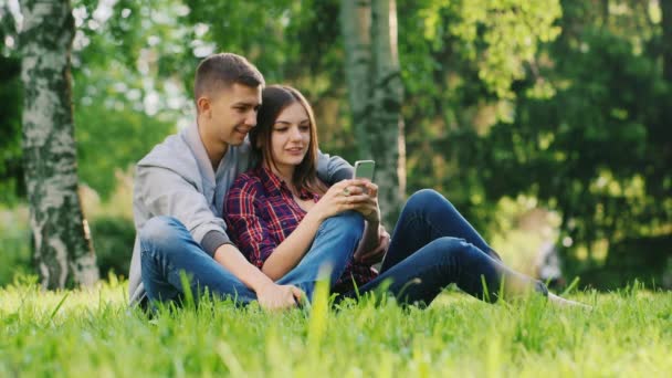 Pareja joven descansando juntos en el parque, jugar en su teléfono, sonriendo . — Vídeo de stock