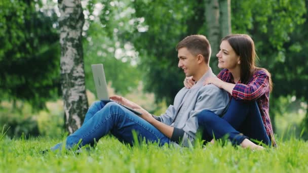 Zaczynając pracę na laptopie. Mężczyzna otwiera ekran laptopa, siedząc na trawie w parku, Kobieta go przytulanie — Wideo stockowe