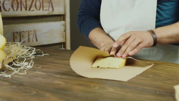 Un vendedor apuesto en el trabajo. Empaqueta un trozo de queso, luego sonríe a la cámara — Vídeos de Stock