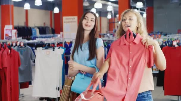 Две привлекательные женщины примеряют платья и сумки перед зеркалом в большом магазине одежды — стоковое видео