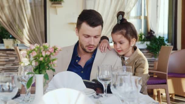 Vader en dochter kocht een diner in een restaurant. Paus houdt de menu, het meisje permanent naast en knuffelen papa — Stockvideo
