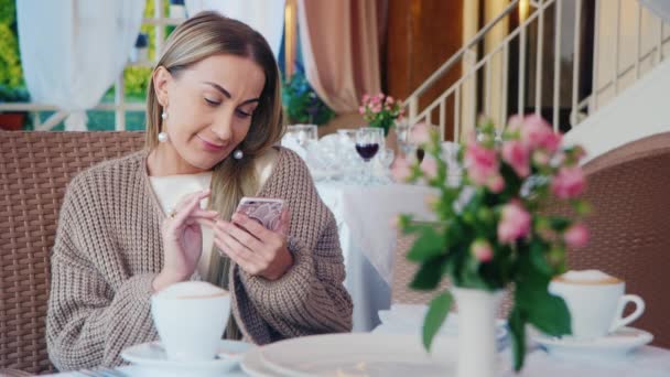 Elegante mujer atractiva utiliza un teléfono inteligente. Sentado en la terraza de verano en la cafetería — Vídeos de Stock