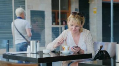 Bir kafede cam kenarında oturan genç kadın. Kahve içme bir cep telefonu üzerinde yazıyor. Dışarıda, insanlar yürümek