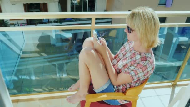 Mujer joven sentada en el balcón de una silla, usando un teléfono móvil. Está escribiendo texto se ve — Vídeo de stock
